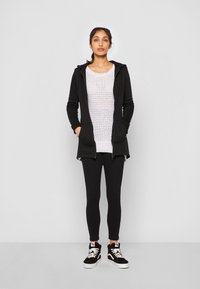 Crna zip-up hoodie sa džepovima, svijetlo ružičasti pleteni top, crne tajice i crne visoke tenisice s bijelim detaljima.