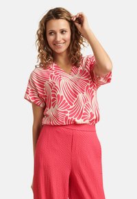 Short-sleeve pink blouse with white leaf pattern, v-neckline, and loose fit, paired with textured pink high-waisted trousers.