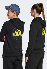 Felpa nera con un logo grafico giallo sul retro, realizzata in tessuto morbido. Abbinata a pantaloni della tuta neri coordinati, con polsini a costine.