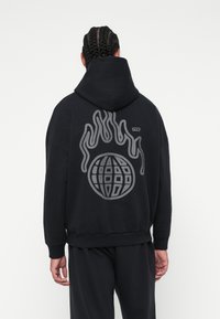 Μαύρη hoodie με χαλαρή εφαρμογή, διαθέτοντας μια μεγάλη γκρίζα γραφική αναπαράσταση μιας υδρογείου περιτριγυρισμένης από στυλιζαρισμένες φλόγες στην πλάτη. Μαλακό, υφασμάτινο υλικό.
