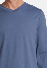 Mann in einem langärmligen blauen V-Ausschnitt-Shirt, gekürzt, um den Oberkörper und die Schultern vor einem weißen Hintergrund zu zeigen.