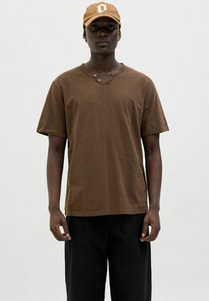Camiseta de algodón marrón con mangas cortas y cuello en V, con dos botones en el escote. Combinada con pantalones holgados negros.