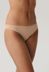 Beżowe, bezszwowe majtki wykonane z miękkiego materiału; niskosiedzący krój z minimalnym zakryciem i gładkimi brzegami. Brak widocznych wzorów lub elementów metalowych.