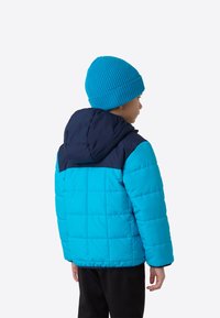 Niebieska pikowana kurtka z granatowymi akcentami, wyposażona w kaptur i pikowaną fakturę. Noszona z dopasowaną prążkowaną czapką. Poniżej widoczne czarne spodnie.