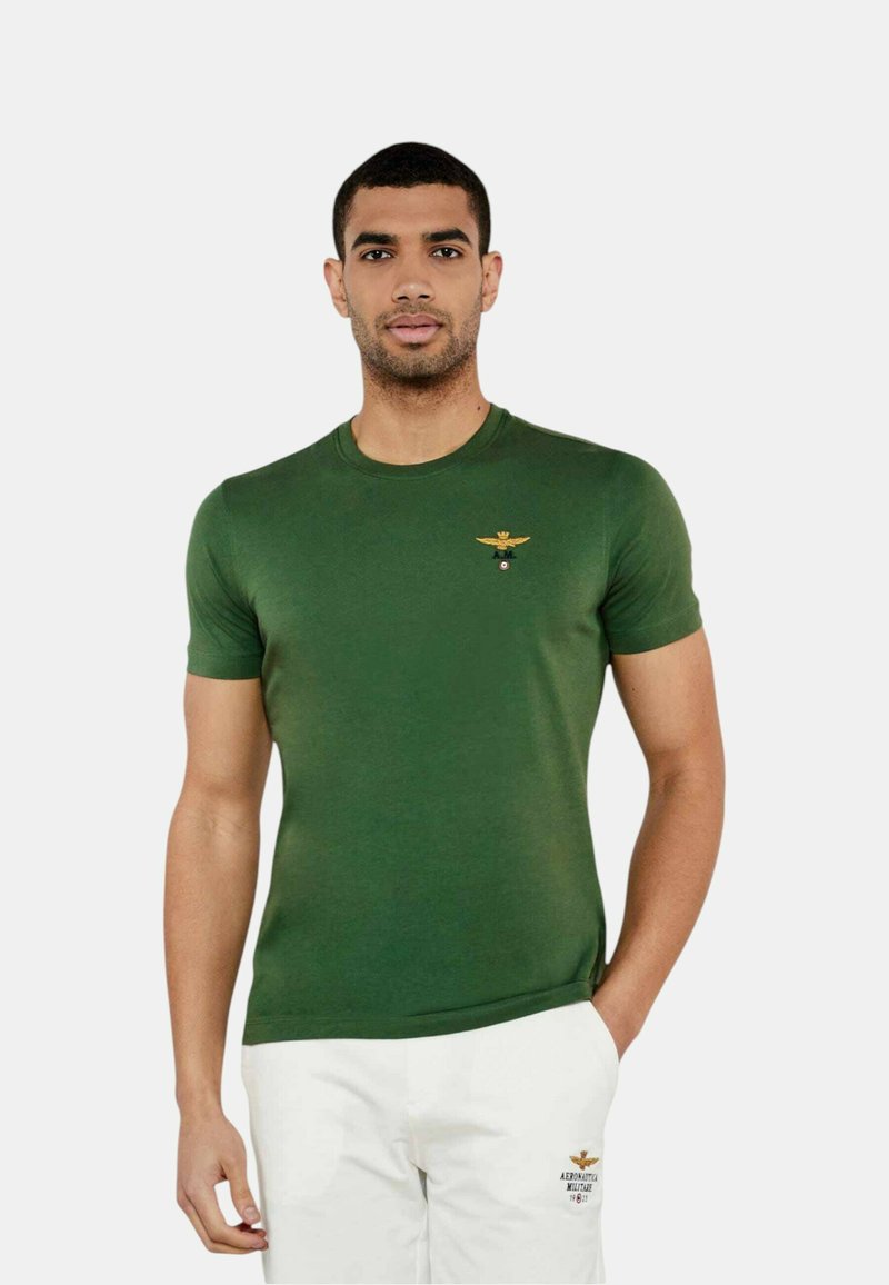 Camiseta verde de manga corta hecha de una mezcla de algodón, con un escote redondo y un pequeño emblema alado en oro y negro en el pecho.