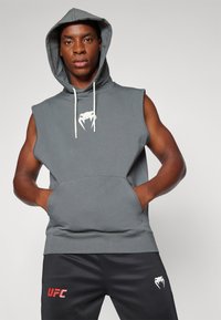 Sivý bezrukávový mikina s predným vreckom a bielym logom. Skombinovaná s čiernymi nohavicami s červeným potlačou "UFC". Hladká bavlnená látka.
