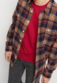 Chemise en flanelle à carreaux bruns et bleus sur un pull rouge. La chemise a un devant à boutons et une poche sur la poitrine gauche. Un jeans sombre est visible.