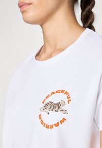 Hvit bomulls T-skjorte med en rund grafikk som har en sprettende tiger og ordene "FREDLIG KRIGER" i oransje og brunt.