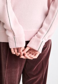 Světle růžový svetr s žebrovanými manžetami a kontrastním zeleným pruhem podél rukávů, spárovaný s měkkými, burgundskými sametovými kalhotami.