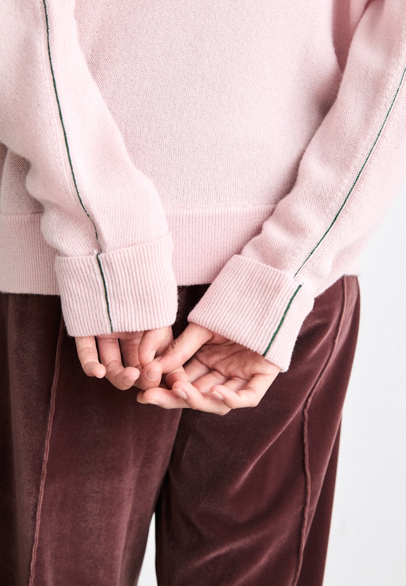 Helles rosa Sweatshirt mit gerippten Manschetten und einem kontrastierenden grünen Streifen entlang der Ärmel, kombiniert mit weichen, burgunderfarbenen Samthosen.