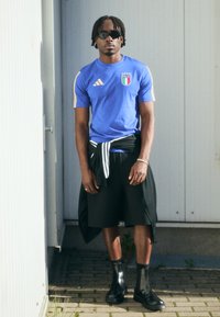 Zils Adidas krekls ar Itālijas karogu, melni šorti ar svītrainu jostu un spīdīgas melnas potītes kurpes. Saulesbrilles piešķir izskanam noslēgumu.