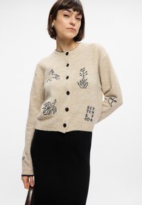 Κρεμ μακριά ζακέτα με πλέξιμο που διαθέτει μαύρες κεντητές σχεδιάσεις φυτών και ζώων, με κουμπιά στο μπροστινό μέρος και ριμπ στεφάνι. Μαύρη φούστα από κάτω.