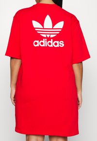 Czerwona koszulowa sukienka z bawełny o luźnym kroju, z widocznym białym logo Adidas i wzorem trzech pasków na plecach. Krótkie rękawy.