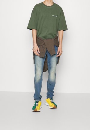 T-shirt surdimensionné vert olive avec texte blanc, sweat-shirt marron noué à la taille, jeans bleu clair et baskets multicolores.