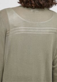 Olivengrøn strikket cardigan med tekstureret ryg, der har tre vandrette rækker med hulmønster og lukning med en enkelt knap ved halsen.