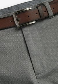 Sivi uzorčasti materijal hlača s smešnim kožnim remenom koji ima srebrnu metalnu kopču, prikazujući detaljno šivanje i teksturu.