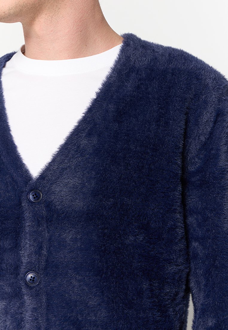 Cardigan en velours bleu marine avec un design en V, comportant deux boutons et une texture douce, superposé à un T-shirt blanc.