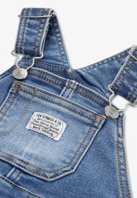 Salopetă din denim albastru deschis cu accesorii argintii. Dispune de bretele reglabile, cusături portocalii și o etichetă pe buzunarul din spate.