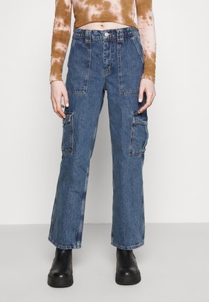 Personne portant un pantalon cargo en denim bleu taille haute avec de grandes poches avant et latérales, des bottes noires et un haut court marron tie-dye à manches longues.