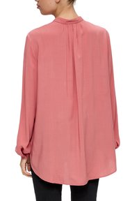 Langwerpige, roze blouse met een zachte, gladde textuur. Heeft een mandarin kraag en plooien aan de achterkant, wat een ontspannen silhouet creëert.