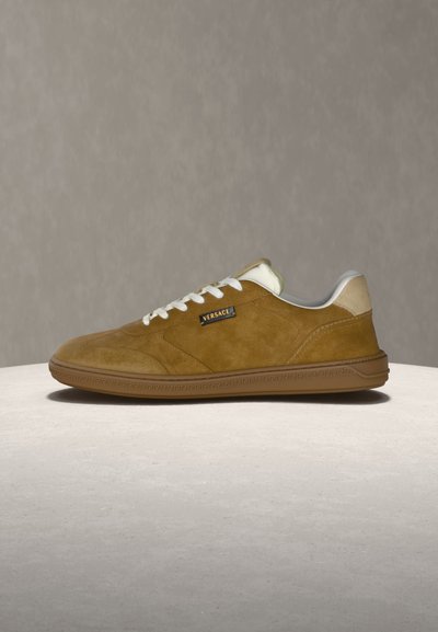 Baskets basses en daim marron avec lacets blancs, détail talon beige et logo Versace sur le côté, présentées sur une surface claire.