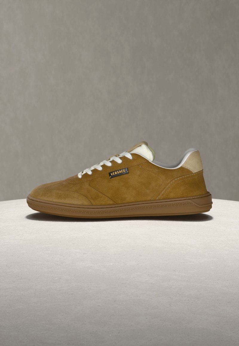 Brauner Wildleder-Sneaker mit flacher Sohle, weißen Schnürsenkeln, beigen Fersendetails und Versace-Logo an der Seite, auf einer hellen Oberfläche präsentiert.