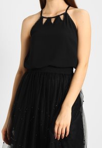 Haut noir à col halter avec découpes triangulaires décoratives et bordure perlé, associé à une jupe en tulle noire ornée de perles dispersées.