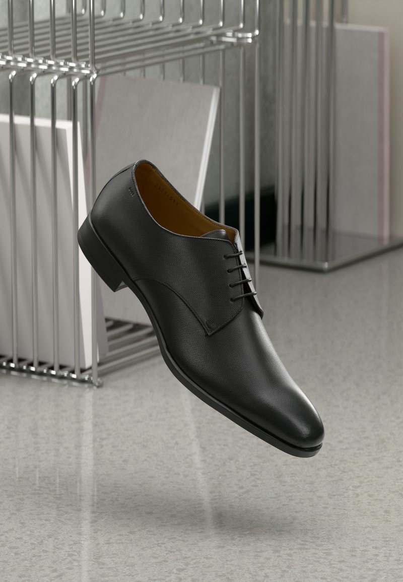 Chaussure habillée en cuir noir avec lacets flottant au-dessus d'un sol gris poli près d'une étagère en fil métallique contenant des livres blancs.