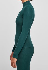 Frau trägt ein tailliertes, tealfarbenes, geripptes Langarmshirt und einen passenden gerippten Rock, steht seitlich vor einem schlichten grauen Hintergrund.