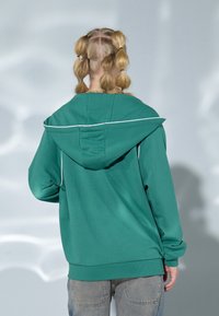 Sudadera con capucha verde con detalles de ribete blanco, que presenta un corte relajado, mangas largas y puños acanalados, vista desde la parte posterior.