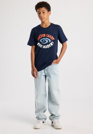 T-shirt bleu marine avec un logo vibrant blanc et orange "GOLDEN COAST SURF ACADEMY." Associé à un jean baggy bleu clair et des baskets blanches.