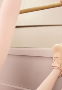 Scarpa da atletica di colore rosa chiaro con tomaia in maglia strutturata e fascetta in velcro, posizionata accanto a una superficie liscia e chiara con uno sfondo neutro.