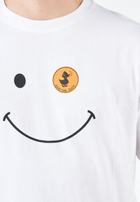 T-shirt bianca in cotone con una grande grafica nera di un sorriso e un'etichetta circolare arancione con un logo di anatra e il testo "SALVA L'ANATRA".
