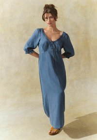 Denim maksi kleita ar puff piedurknēm, dziļu V-veida kaklu un aukliņas detaļu centrā. Mīksta tekstūra gaiši zilā krāsā.