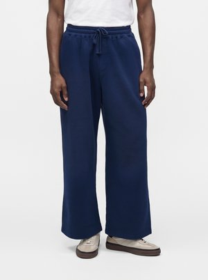 Homme portant un pantalon de jogging large bleu marine avec taille à cordon et des baskets blanches, debout devant un fond clair uni.