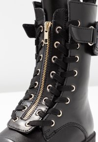 Botte en cuir noir avec fermeture éclair dorée, lacets noirs et œillets. Le design comprend une sangle et un panneau texturé contrastant en haut.