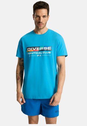 Turkusowa koszulka z krótkim rękawem z wielokolorowym nadrukiem z napisem "DIVERSE NAUTICAL CLUB". Noszona z dopasowanymi niebieskimi szortami.