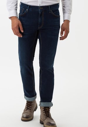 Dark blue denim jeans with a straight fit, featuring light stitching, a button closure, and cuffs rolled up. Paired with brown lace-up boots.