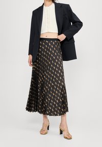 Vrouw in een zwarte blazer over een witte, kortaf, geknoopte top, zwarte gemusterde, geplooide midi-rok en gouden strappy hoge haksandalen.