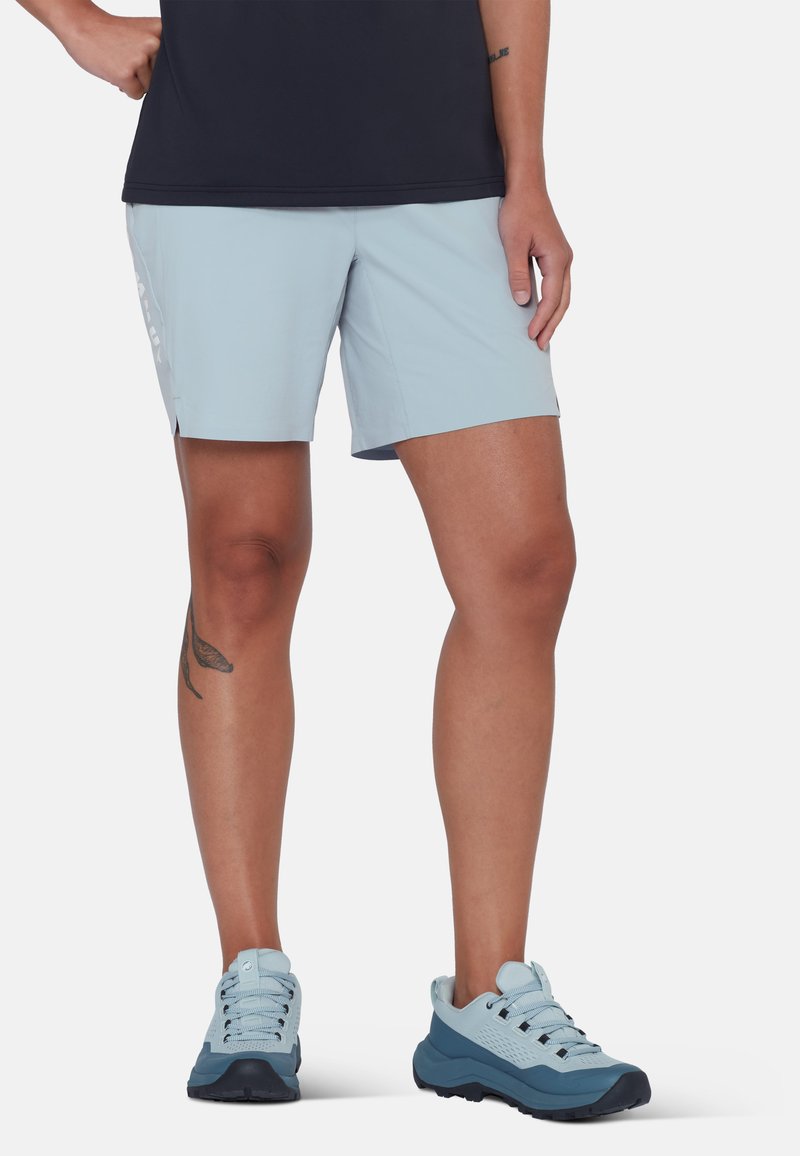 Personne portant un short bleu clair, des baskets assorties bleues et grises, debout avec une main sur la hanche sur fond blanc.