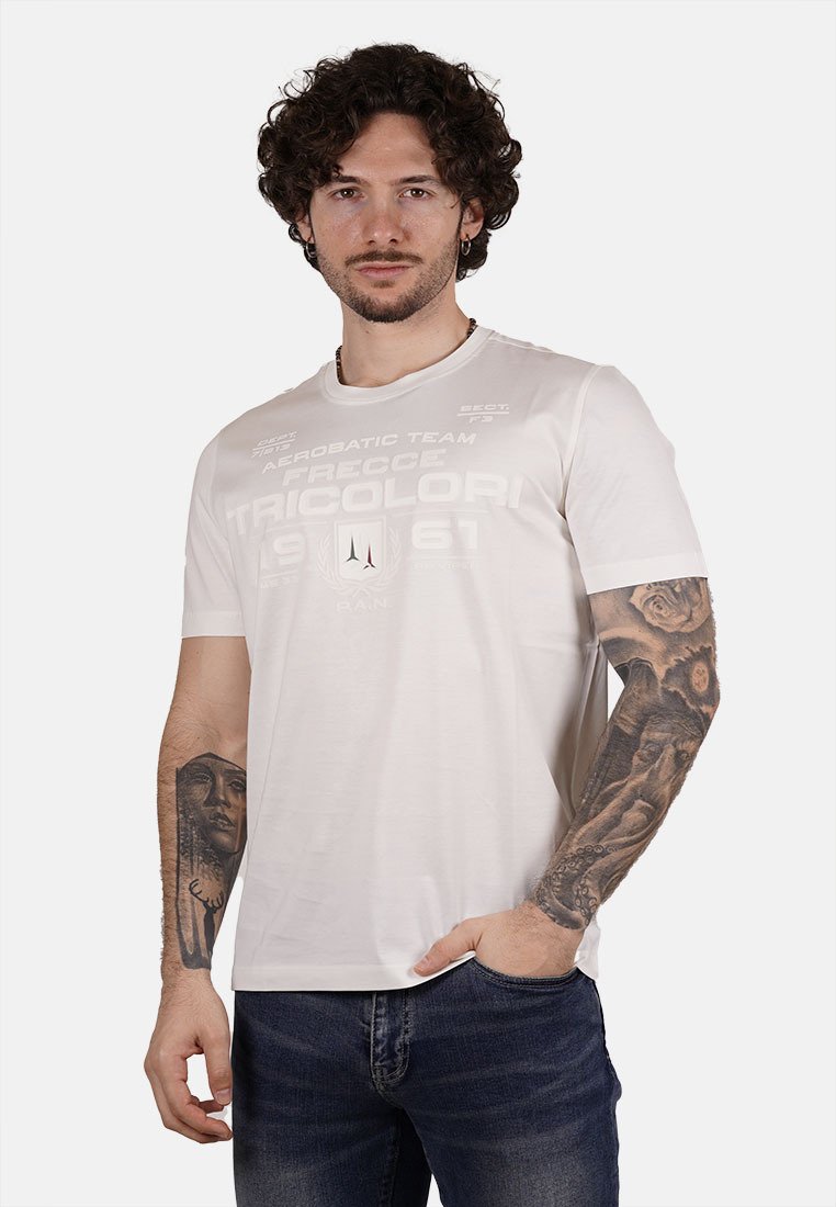 T-shirt bianco a maniche corte con grafica testuale frontale, texture morbida e design a girocollo. Indossato con jeans in denim scuri e tatuaggi visibili.