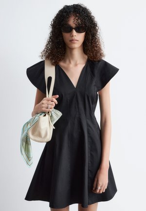 Žena s kučeravými vlasmi nosí čierne šaty so širokými krátkymi rukávmi, čierne slnečné okuliare, drží béžovú kabelku s vzorovanou šatkou na ramene.