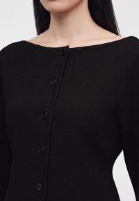Femme portant un cardigan noir ajusté boutonné, avec un large col et des manches longues sur un fond uni.