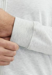 Grå bomulds Sweatshirt med ribbede manchetter. Nærbillede af hånd, der griber om ærmet, hvilket fremhæver den bløde tekstur og det subtile lysmønster.