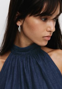 Mörkblå halterneck-top i texturerad tyg, med en samlad halsringning. Bärs med stora silverfärgade hoops, modellens profil synlig.