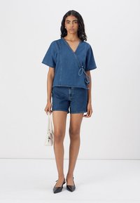 Top in denim con maniche corte, di colore blu scuro, con dettaglio a fiocco in vita, abbinato a pantaloni corti in denim e décolleté nere a punta.
