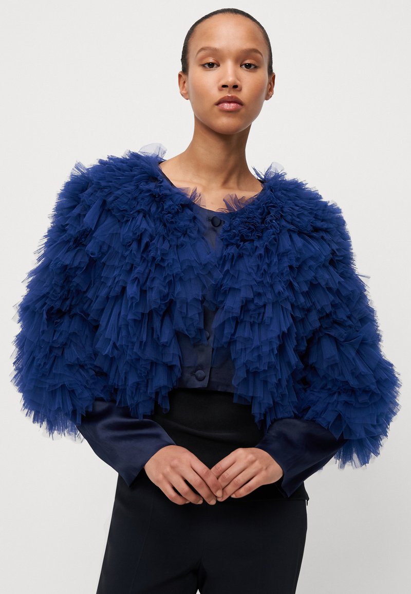 Granatowa krótka kurtka z warstwami tiulowych falbanek, okrągłymi guzikami i gładkim satynowym wykończeniem. Dopasowane czarne spodnie uzupełniają całość.