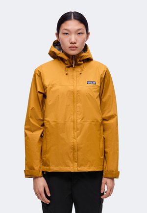Personne portant une veste imperméable jaune moutarde avec capuche, debout face à l'avant sur un fond clair uni.