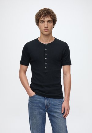 Un homme porte un tee-shirt Henley noir à manches courtes et un jean bleu sur un fond blanc uni.