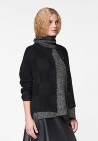 Zwarte gebreide cardigan met een ruitpatroon, ribgebreide manchetten en een ronde halslijn, gelaagd over een donkergrijze coltrui.